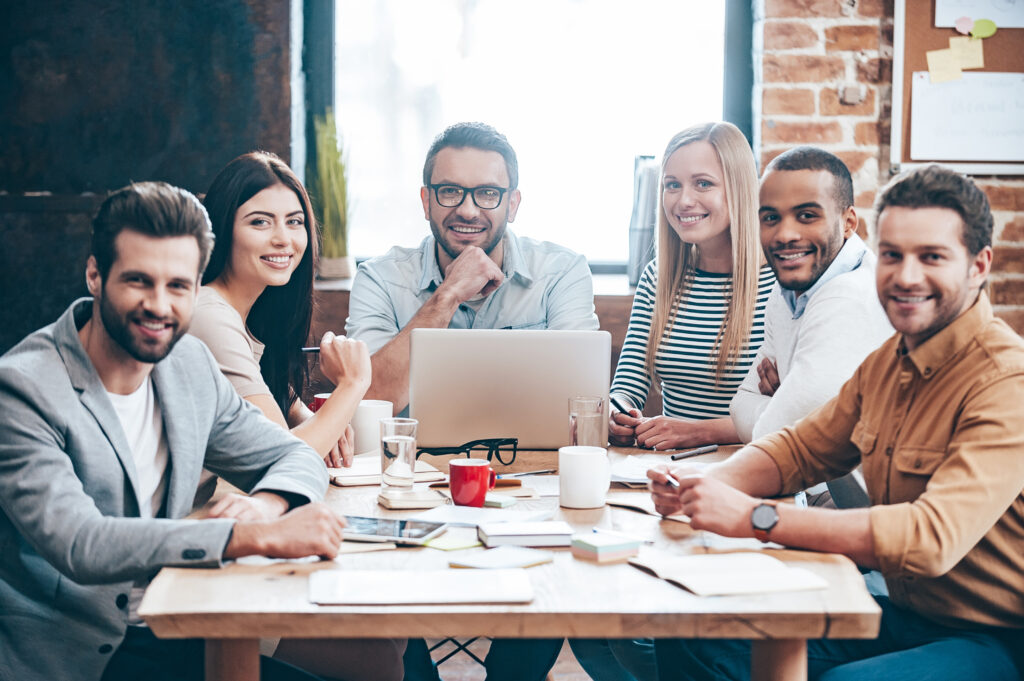 Creative team. Group of six cheerful young people looking at camera with smile while sitting at the table in office